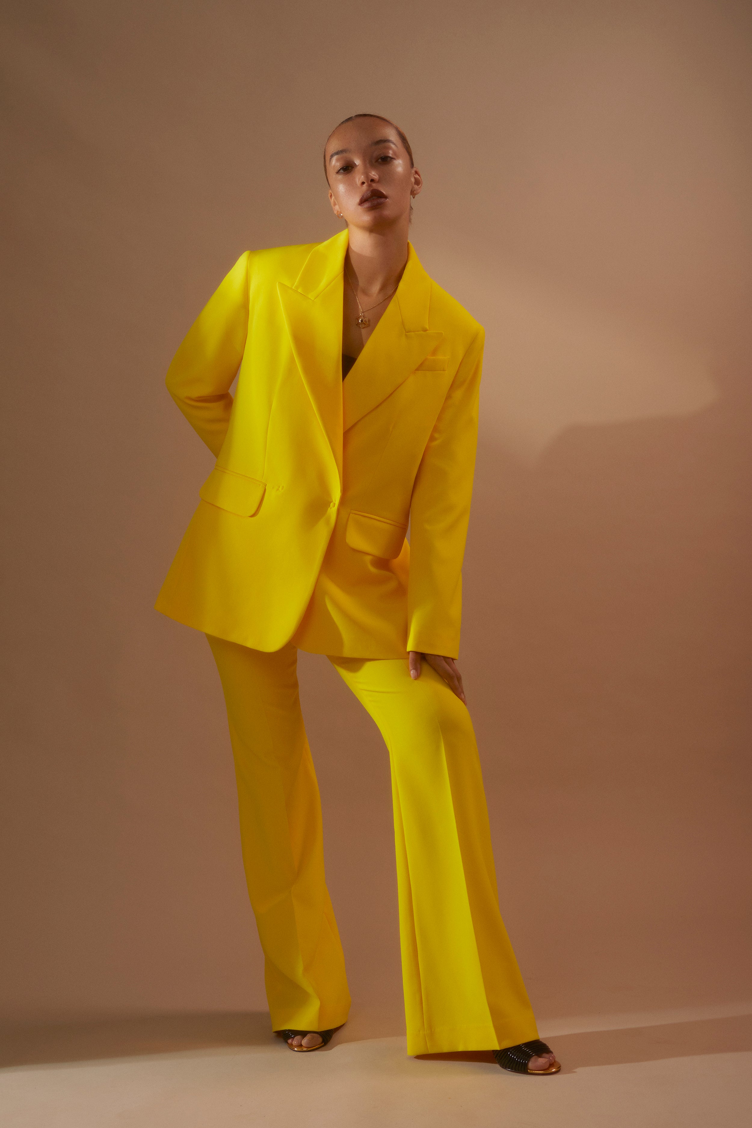 Model wearing yellow suit and Antonia Guise items on a camel background looking to camera