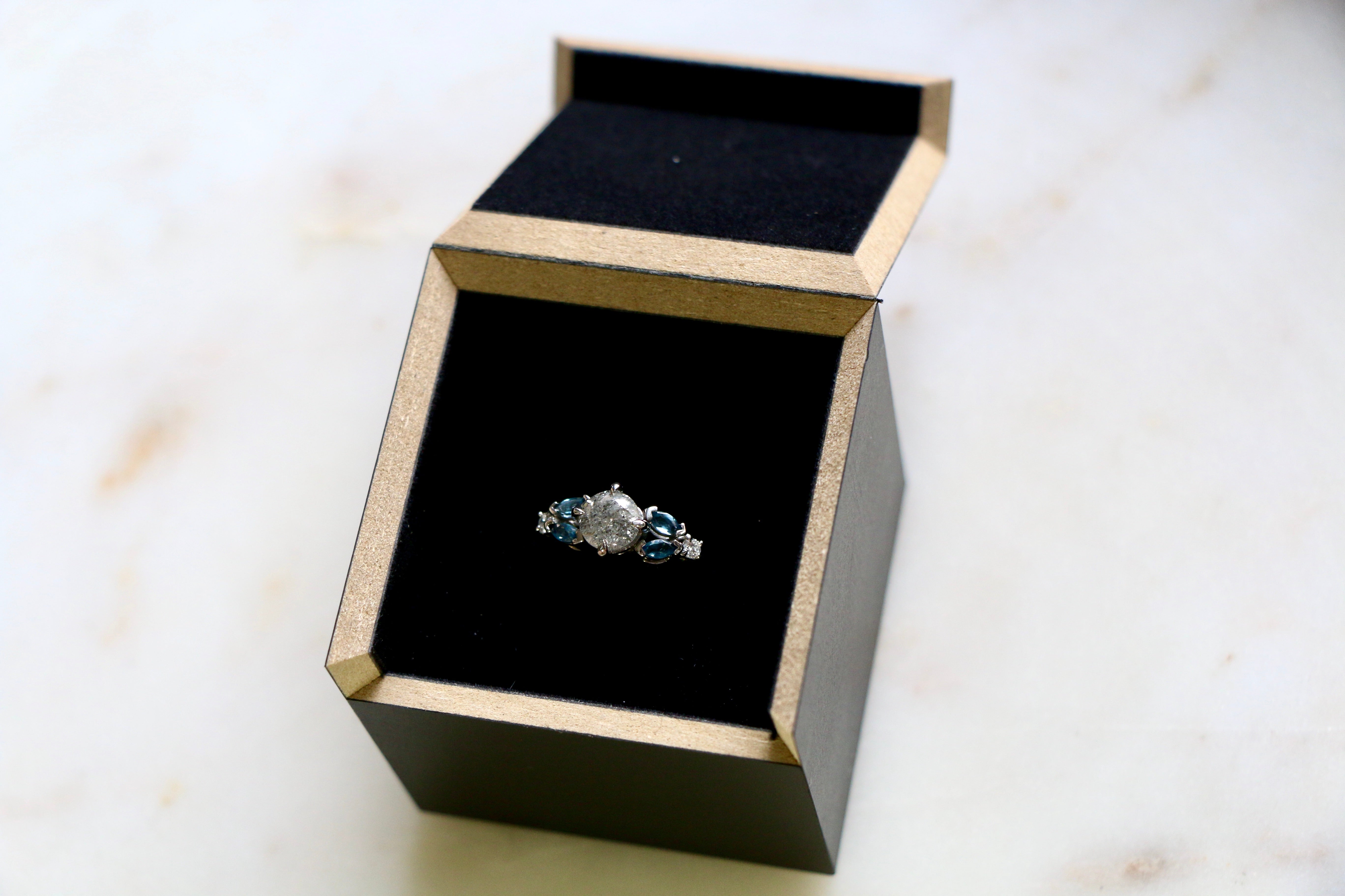 Salt and Pepper diamond engagement ring with flanking marquise topaz stones set in white gold. In it's presentation box on a marble surface.