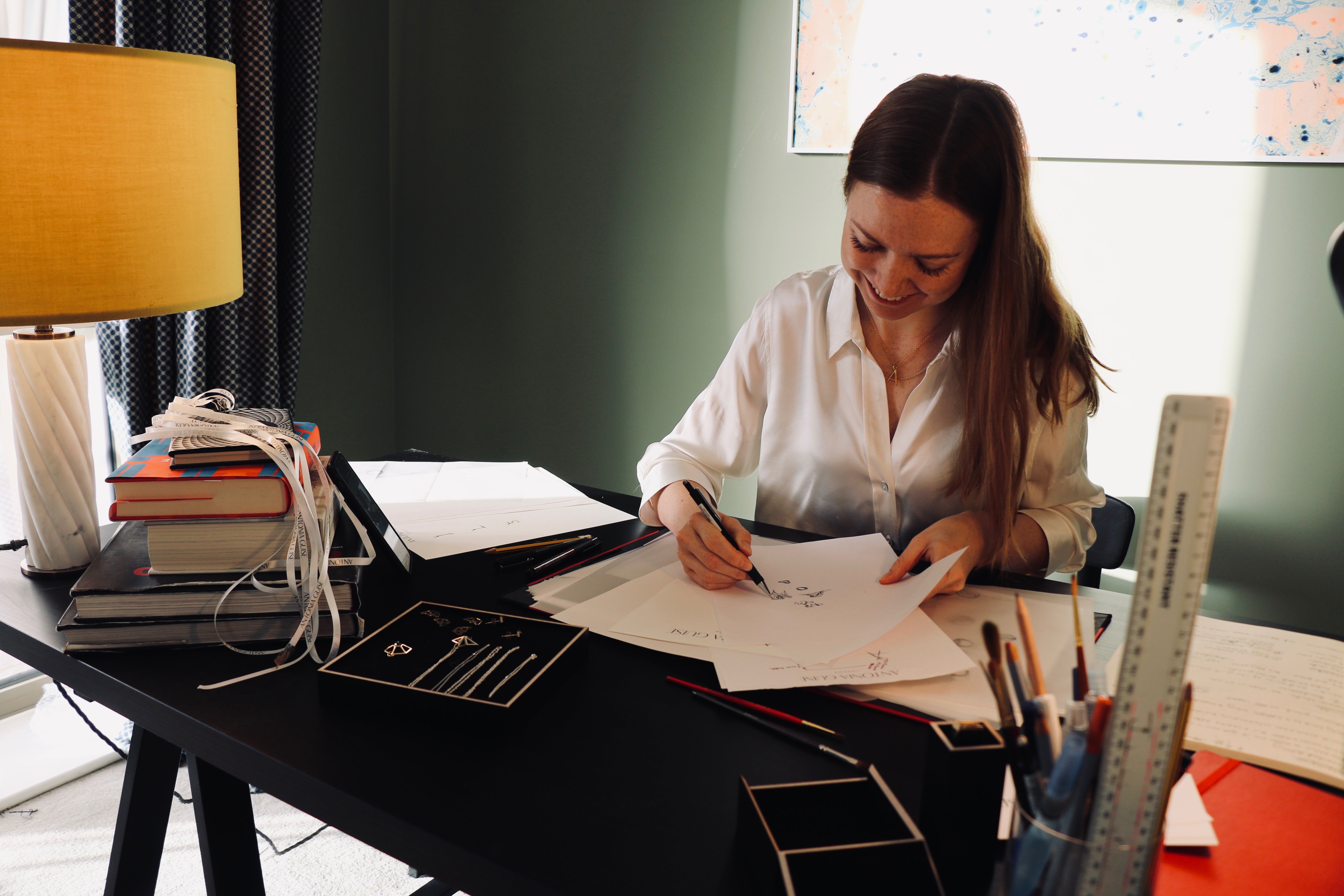 Image of our Founder and Designer Antonia Designing at her desk wearing a white silk shirt and Antonia Guise gold vermeil peices