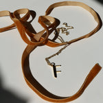 F initial necklace on a white background with shadows and mustard velvet ribbon snaking around it
