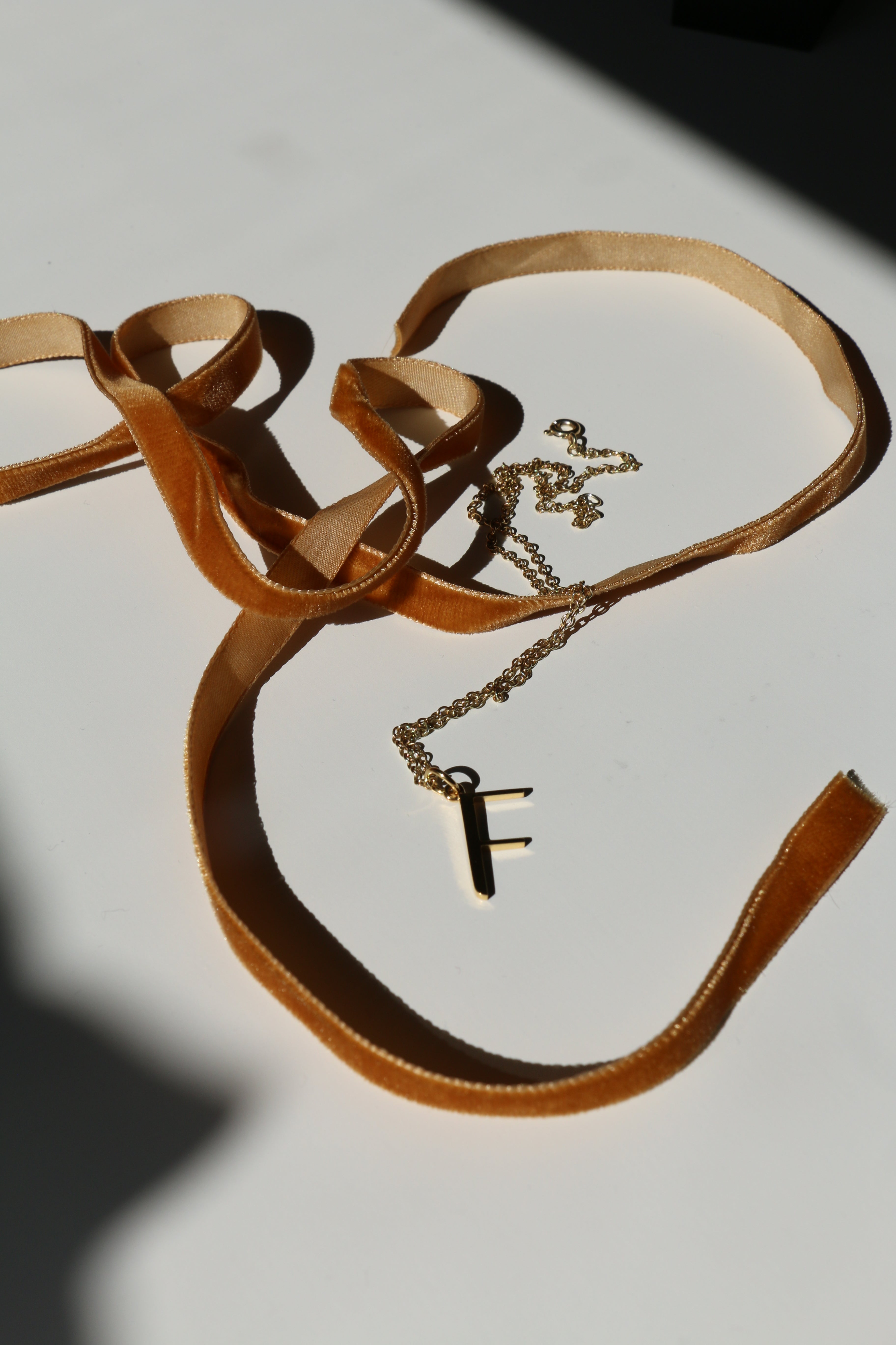 F initial necklace on a white background with shadows and mustard velvet ribbon snaking around it