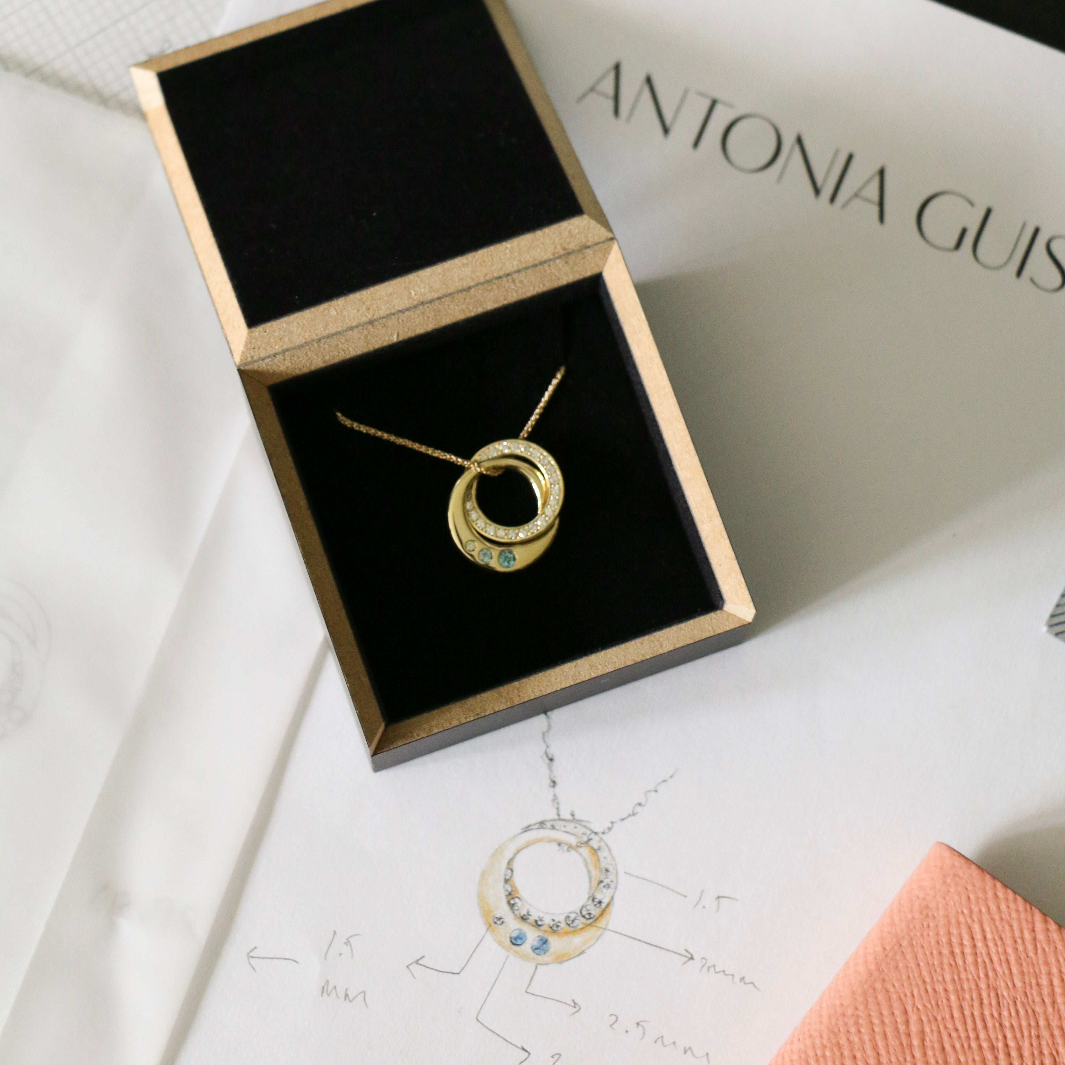 Photo of a bespoke diamond, topaz and yellow gold interlocking ring pendant in its box on top of the original designs and a pink note book