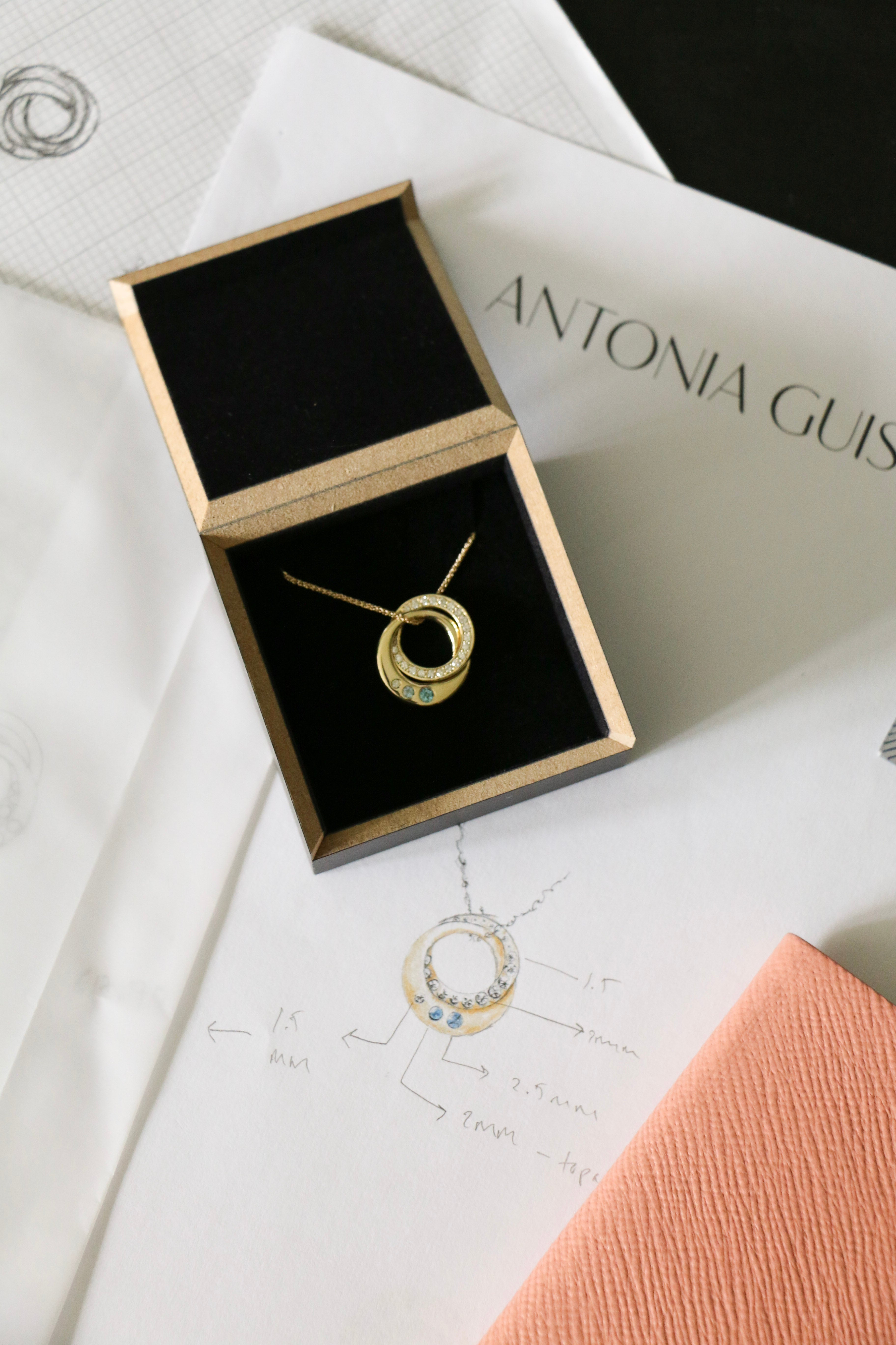 Photo of a bespoke diamond, topaz and yellow gold interlocking ring pendant in its box on top of the original designs and a pink note book