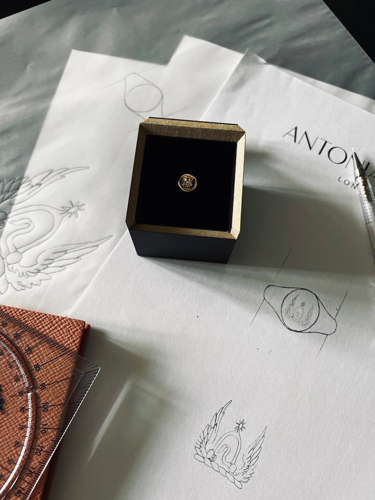 Photo of a bespoke yellow gold signet ring with family crest engraved on it in its box on top of the original designs and a pink note book