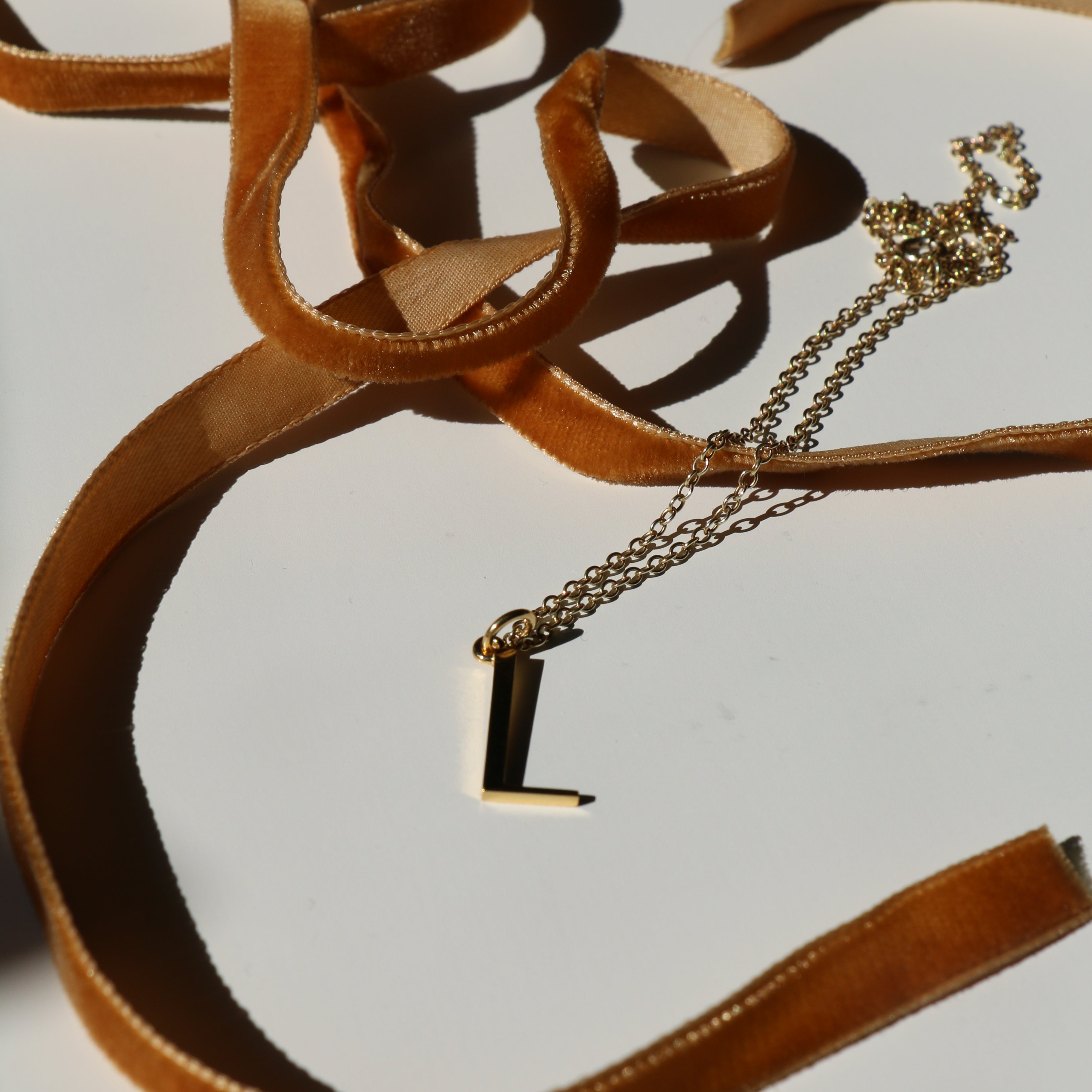 L initial necklace on a white background with shadows and mustard velvet ribbon snaking around it