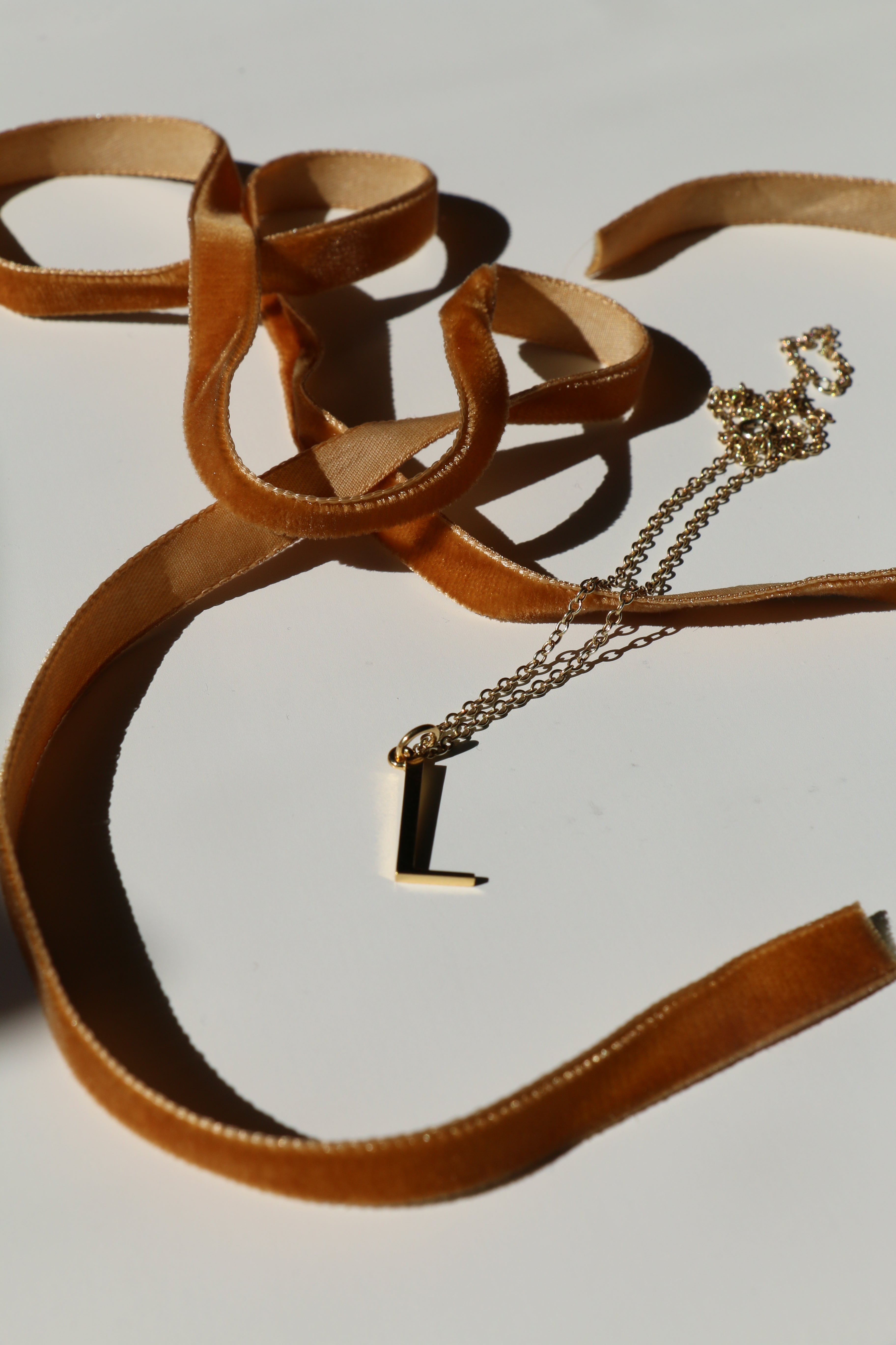 L initial necklace on a white background with shadows and mustard velvet ribbon snaking around it