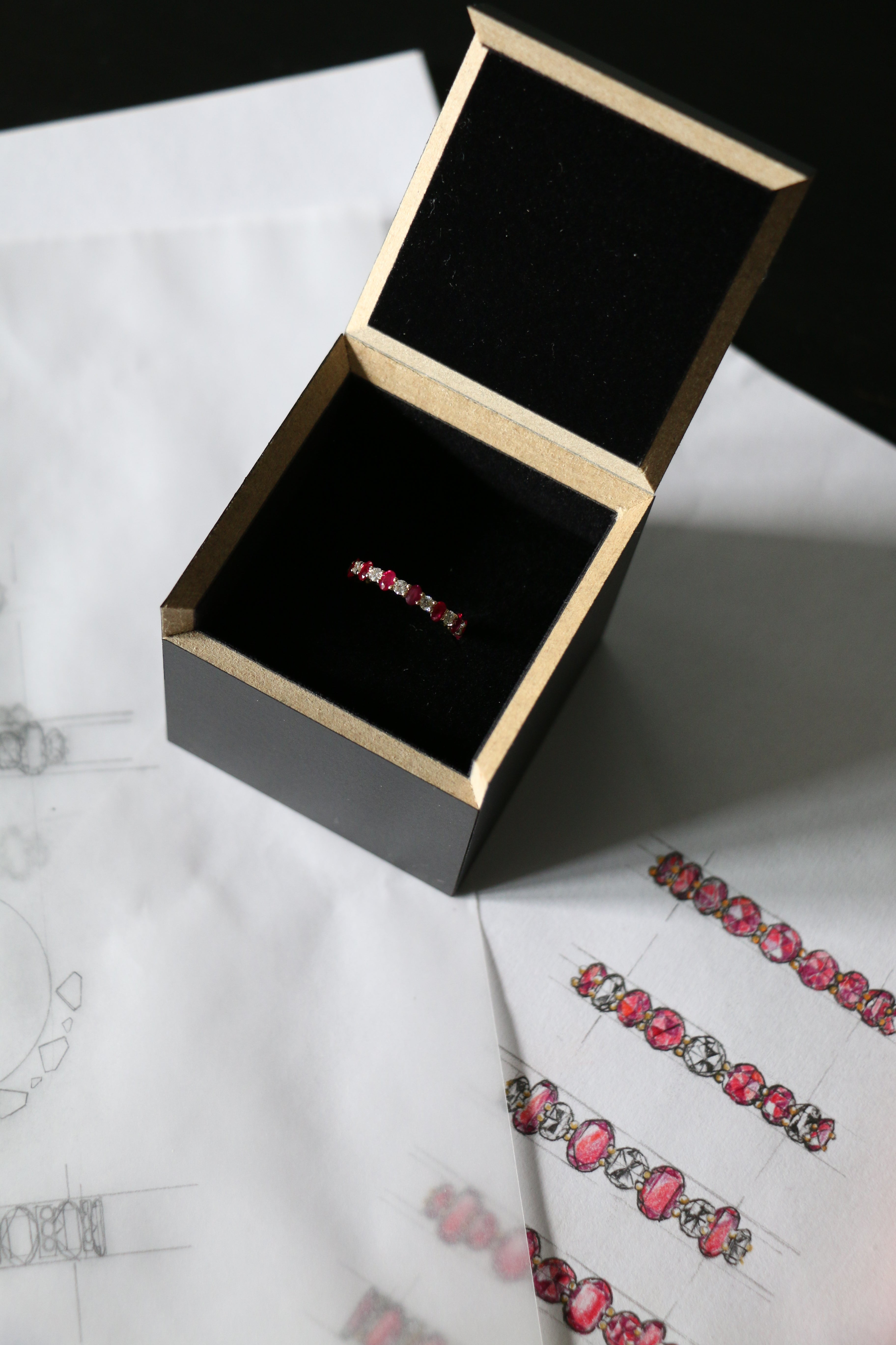 Image of oval Pink Ruby and round Diamond eternity band set in yellow gold in its  black recycled MDF presentation box on top of the original drawings