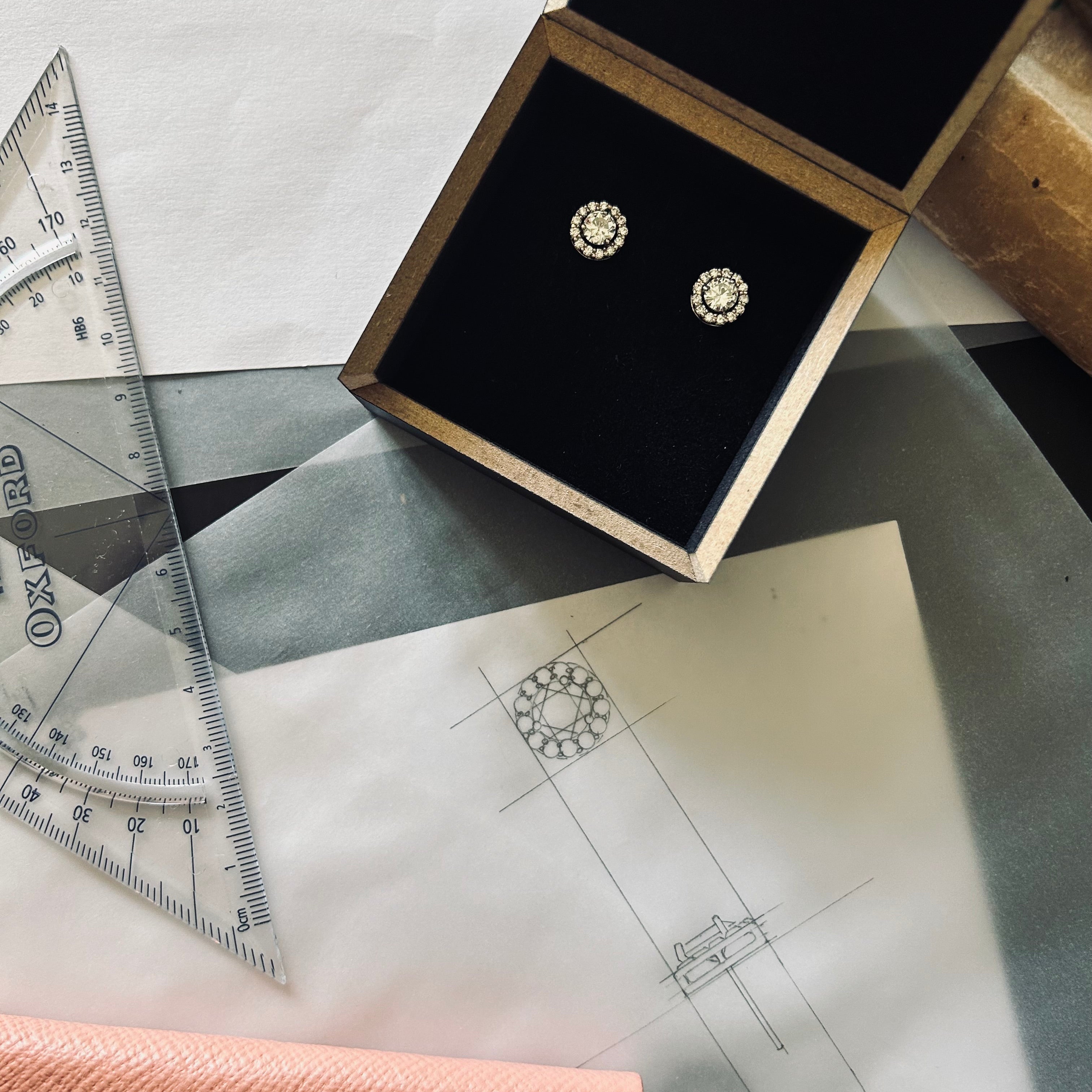 Pair of bespoke repurposed diamond halo studs in their black presentation box on top of the original sketches 