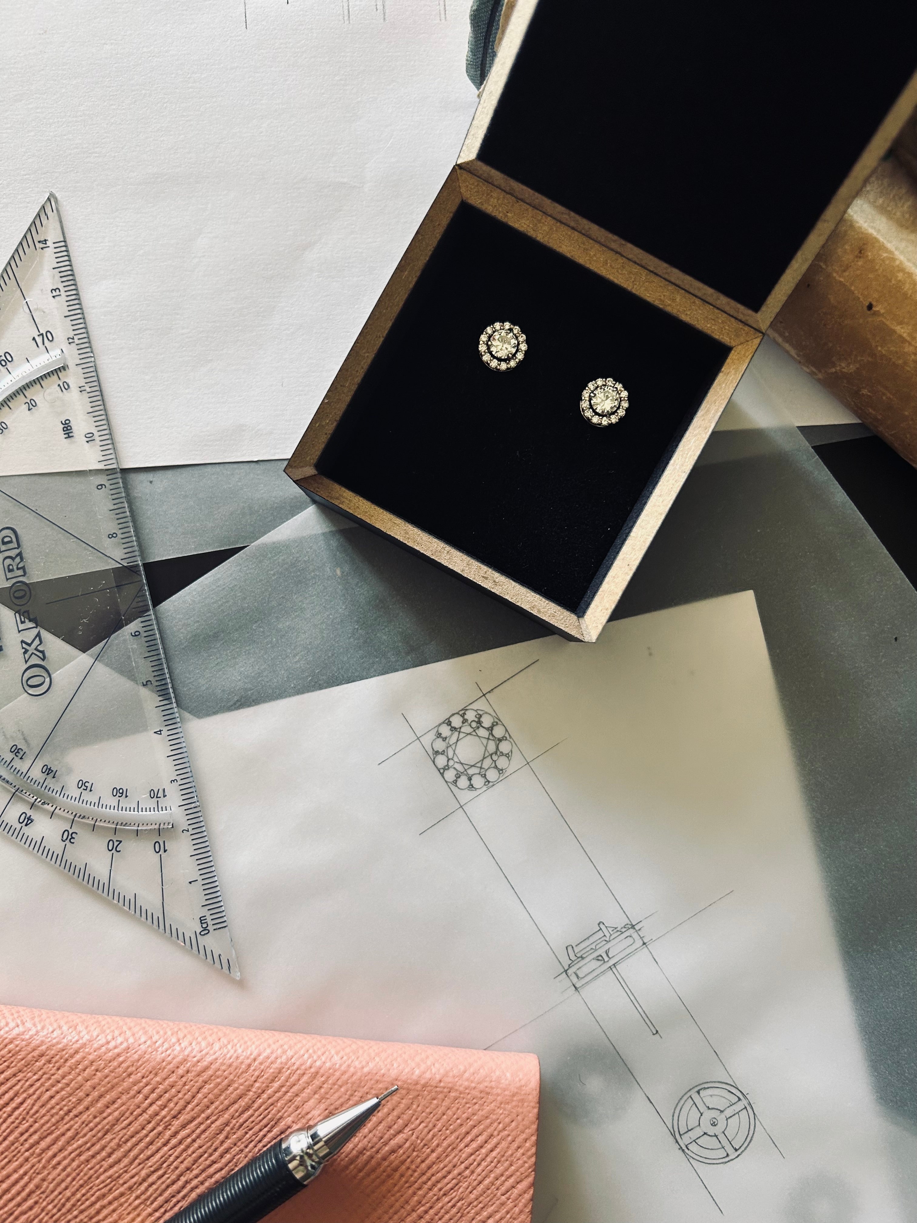 Pair of bespoke repurposed diamond halo studs in their black presentation box on top of the original sketches 